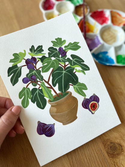 Hand holding a painting of a potted plant with figs on a wooden surface.