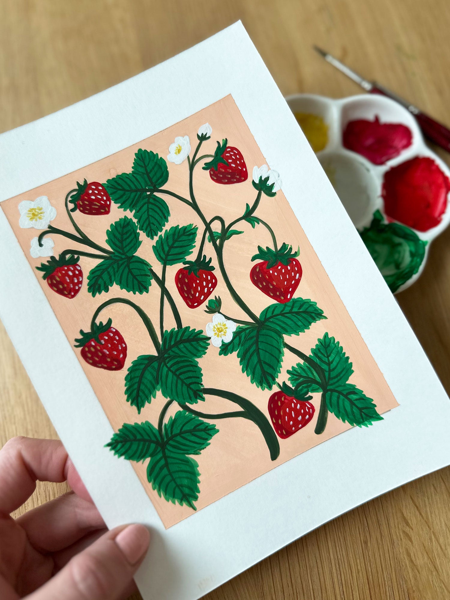 Artistic depiction of strawberries on a peach background held by a hand with watercolor supplies in the background.
